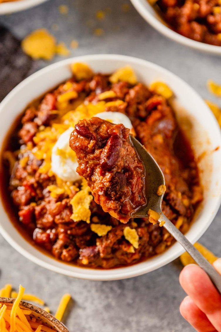 A white bowl of beef chili with a hand in front of the bowl holding a spoonful of chili.