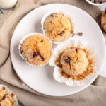 Three Chocolate Chip Muffins on white plate, surrounded by additional muffins