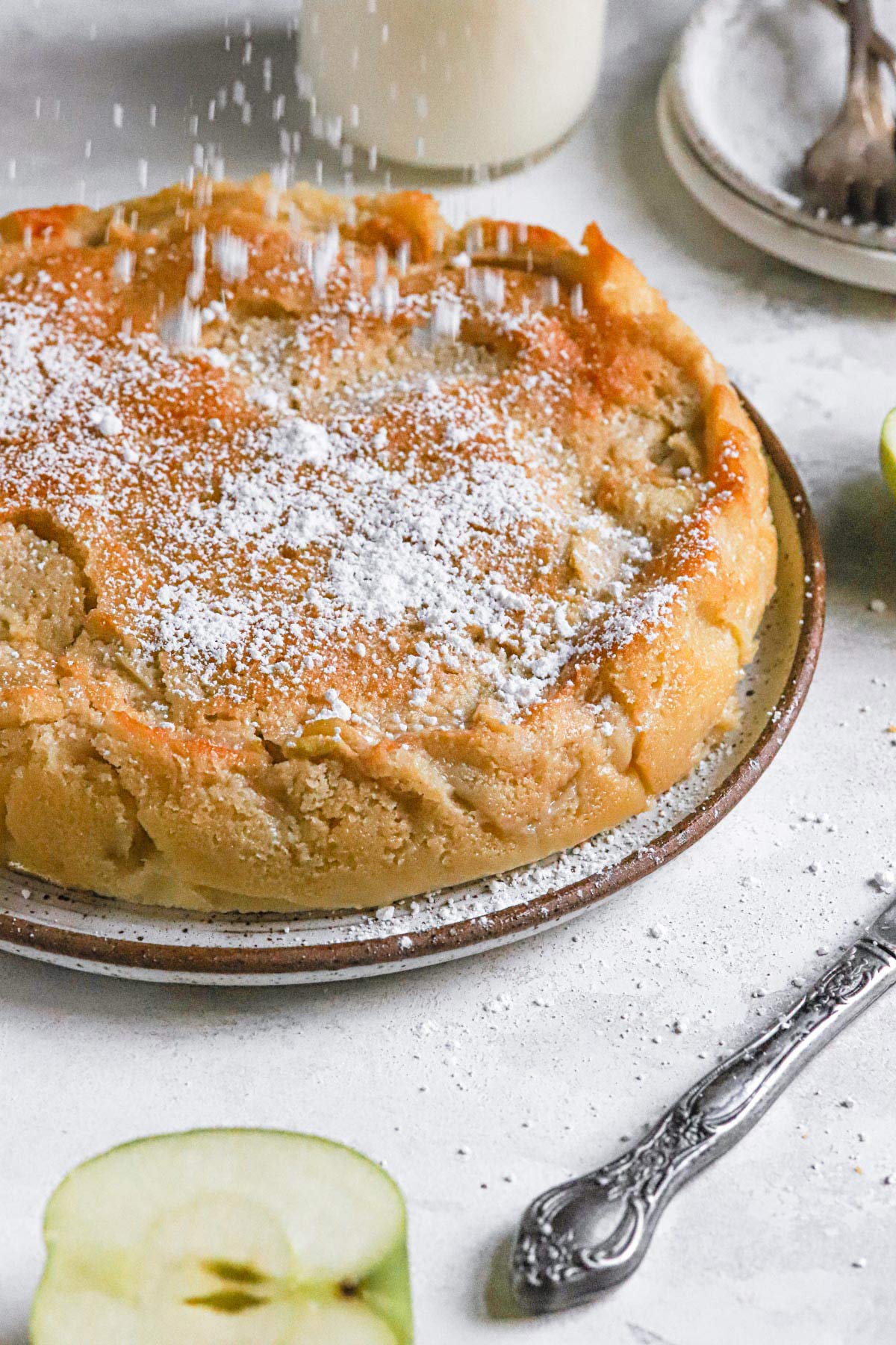French apple cake on a serving plate being dusted with powdered sugar.