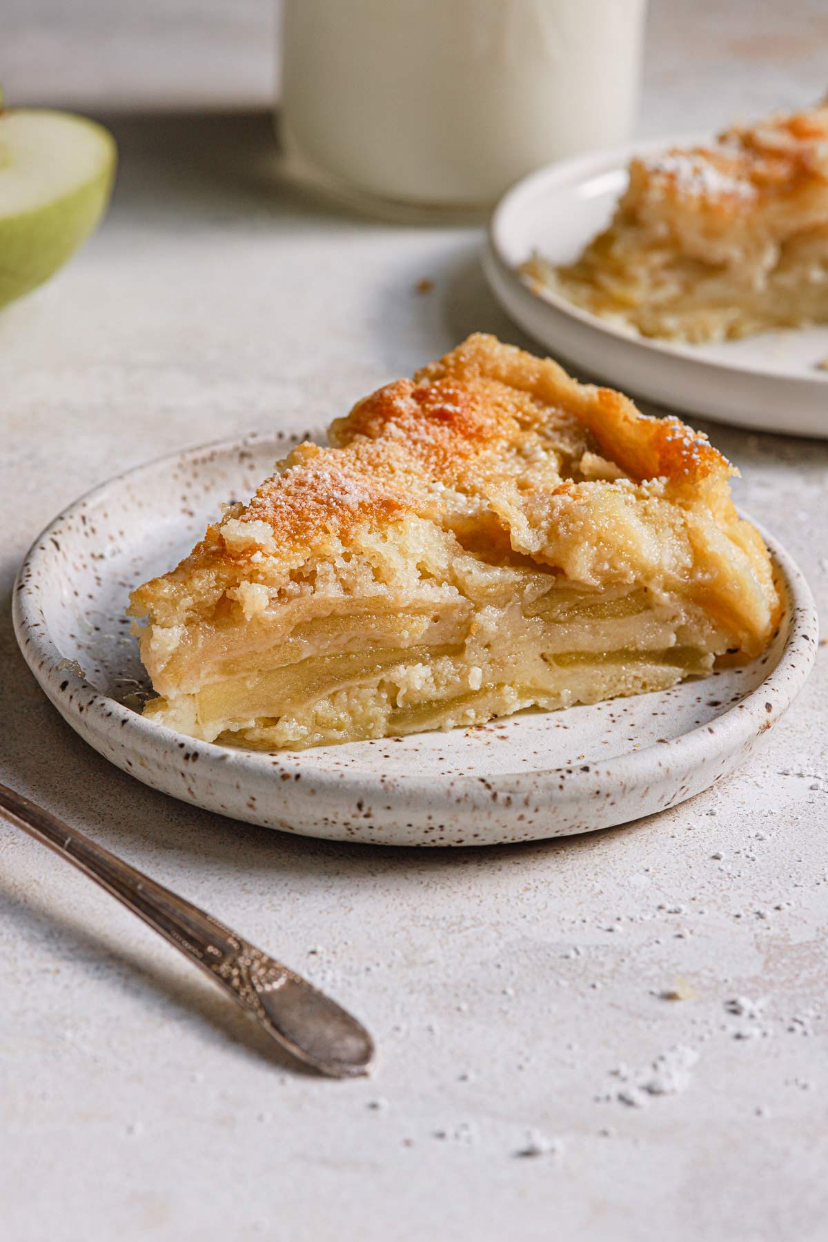 A slice of apple cake on a speckled plate with pieces of a fork and apple in the background.