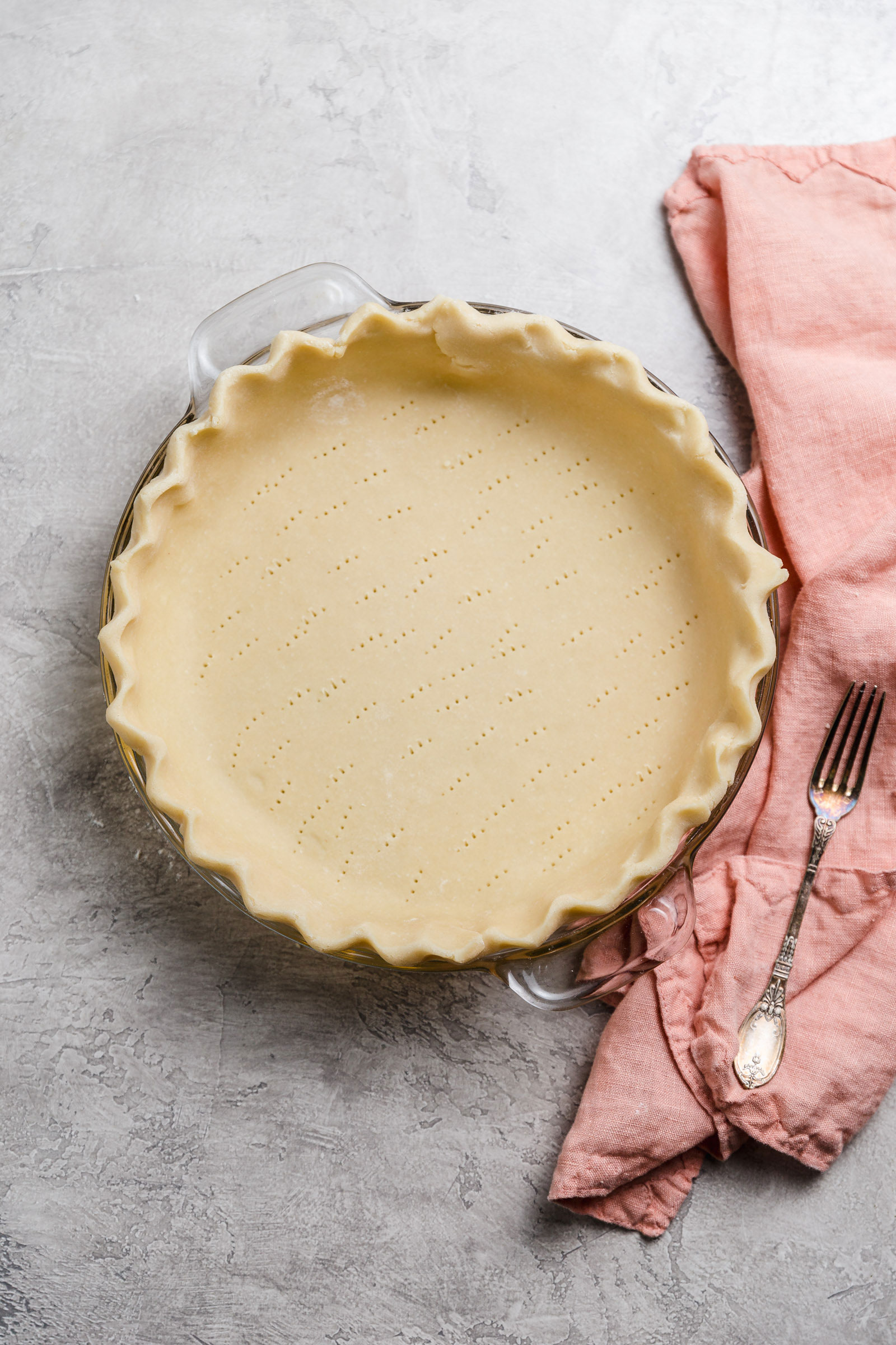 Unbaked pie crust in glass pie plate with fork on a pink kitchen towel next to it.