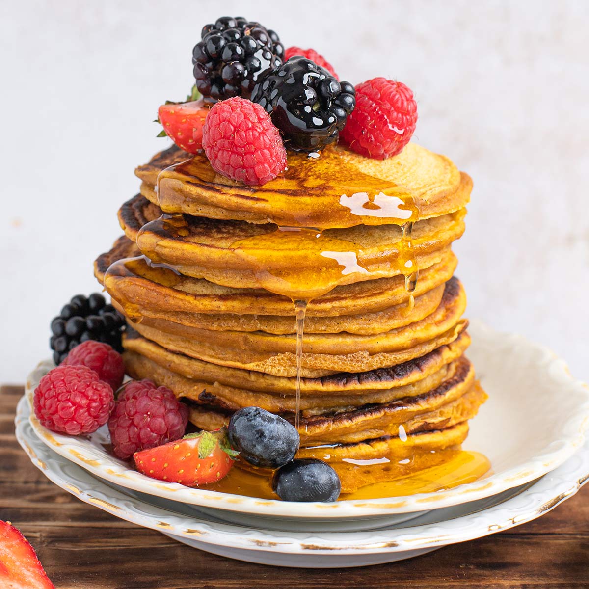 Stack of pumpkin pancakes on plate with syrup dripping down side and berries on top and around bottom.