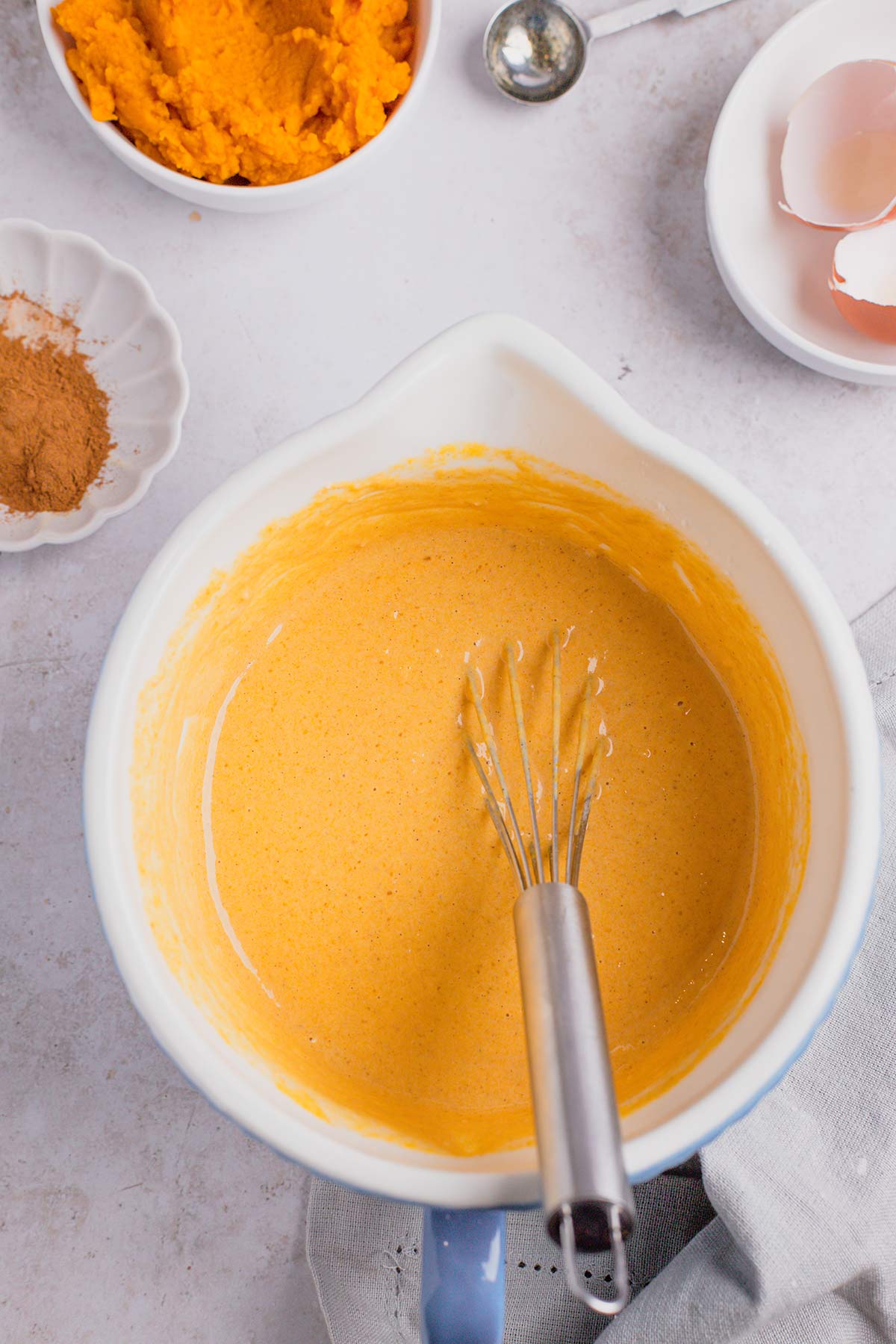 Pumpkin pancake batter in mixing bowl with whisk, with pumpkin puree and spices in bowls surrounding.