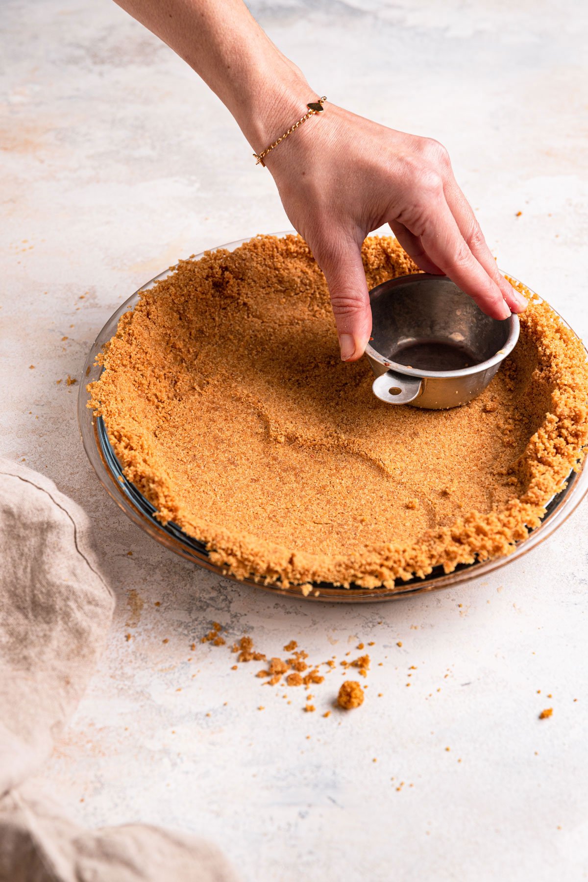 Graham cracker crust being packed into a pie plate using a measuring cup.