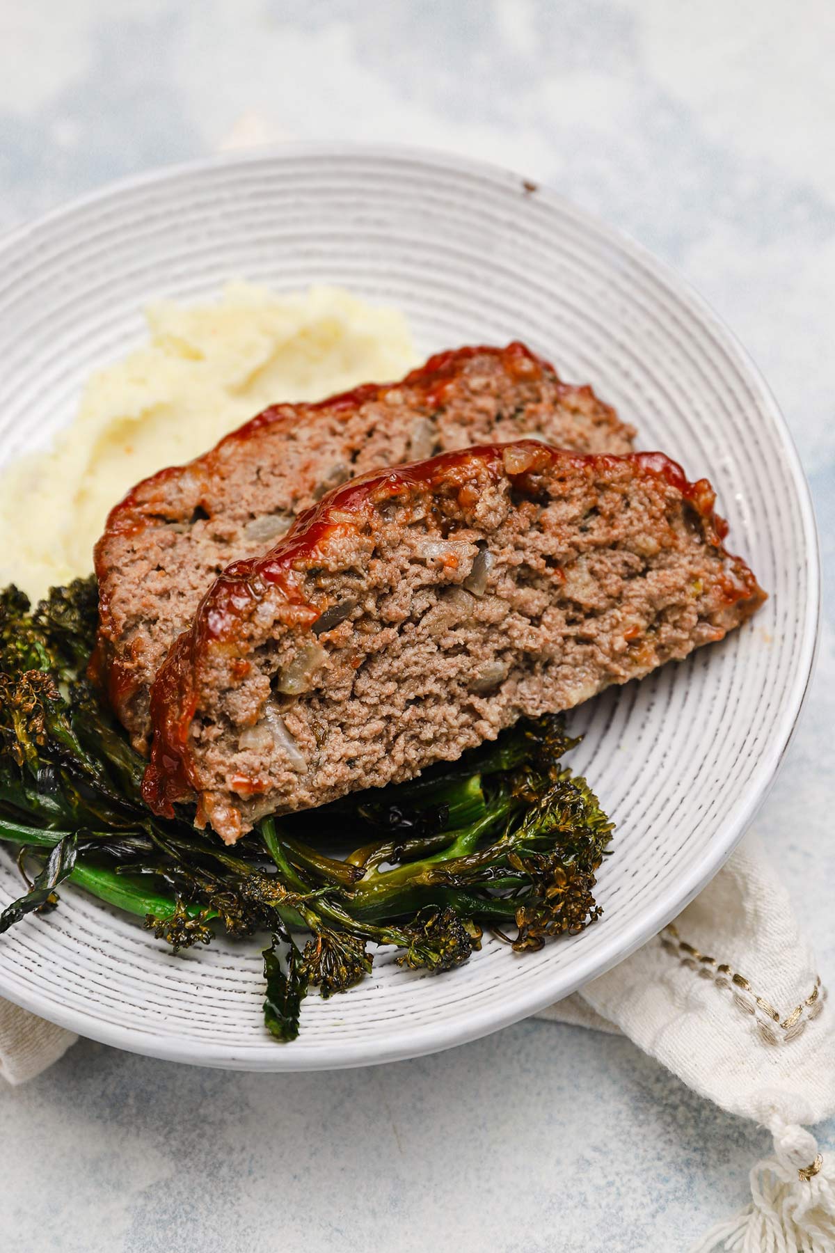 2 slices of meatloaf on a white plate with roasted broccoli and mashed potatoes.