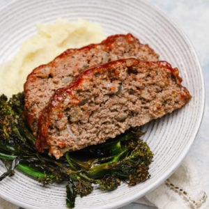 2 slices of meatloaf on a white plate with roasted broccoli and mashed potatoes.