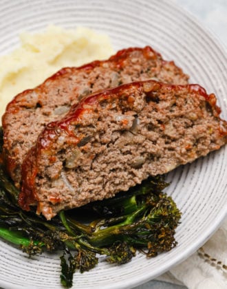 2 slices of meatloaf on a white plate with roasted broccoli and mashed potatoes.