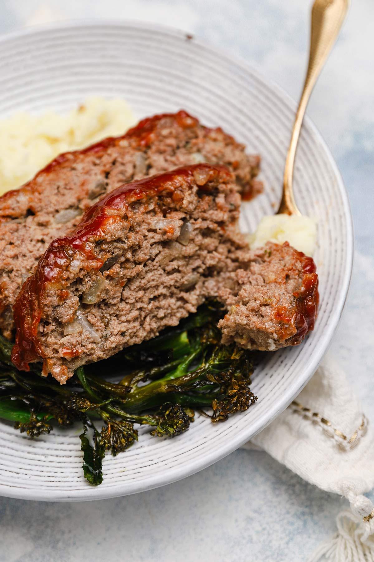2 slices of meatloaf on a white plate with a fork and roasted broccoli and mashed potatoes.