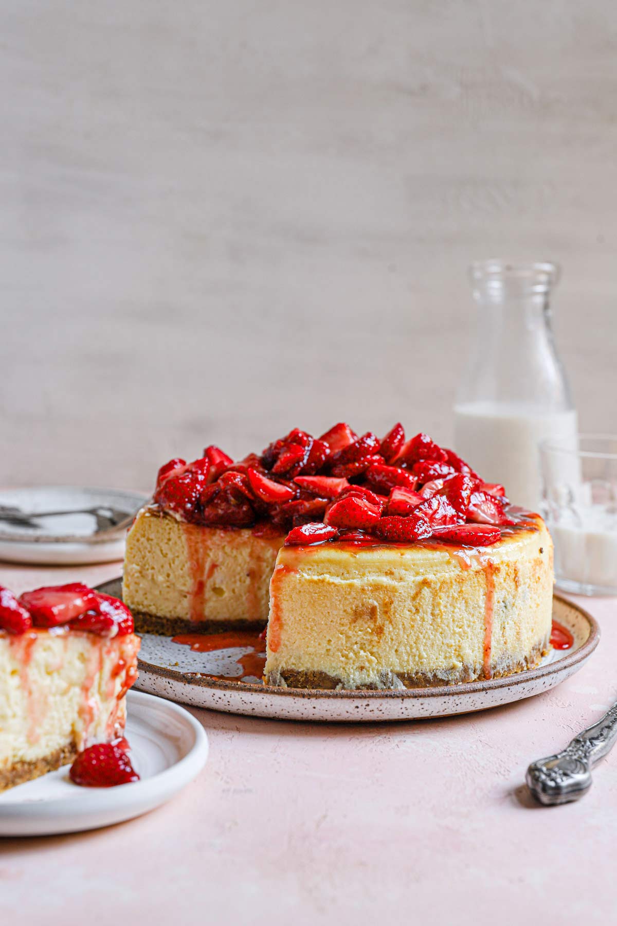 New York cheesecake topped with fresh strawberry sauce on a serving plate with a slice cut out.