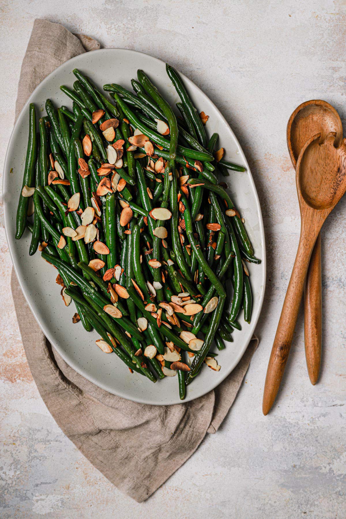 Green beans with toasted sliced almonds in a serving dish with wooden spoons nearby.