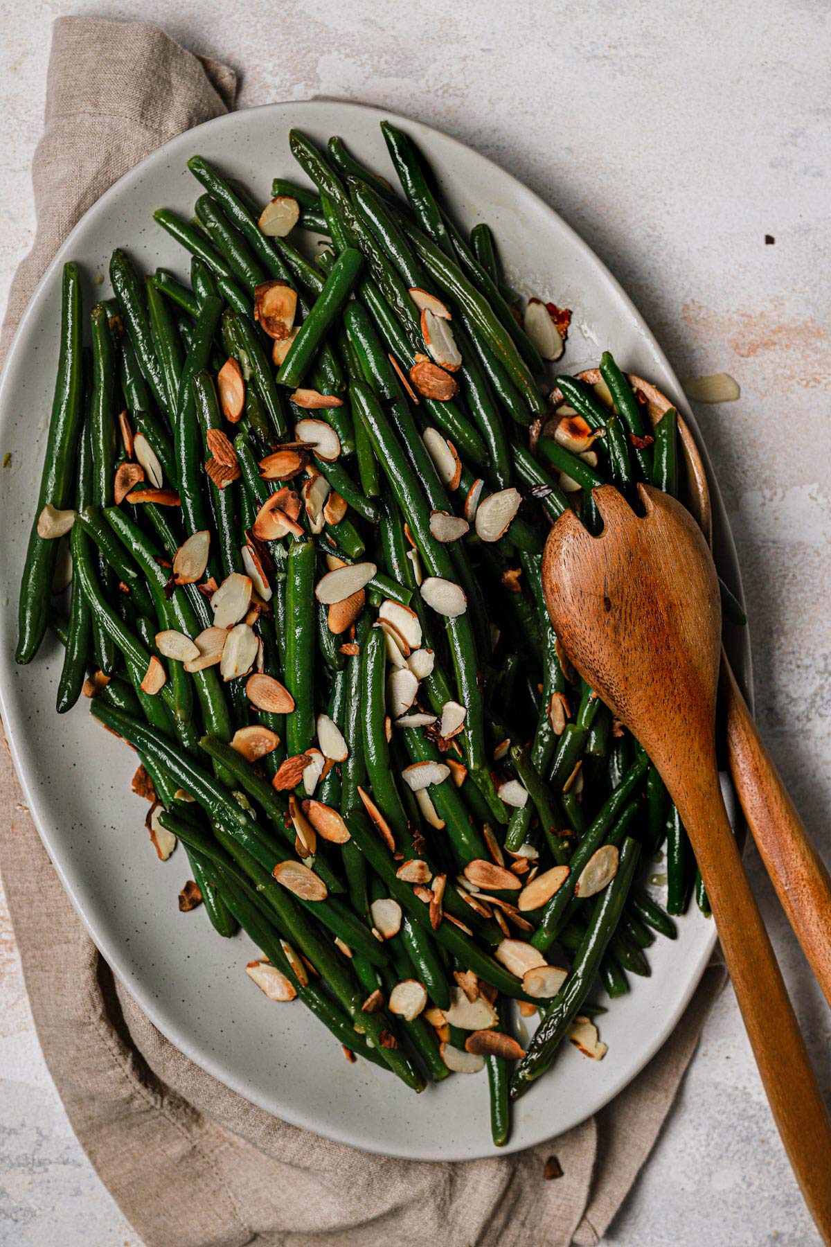 Green beans tossed with toasted sliced almonds in a serving dish with wooden spoons.