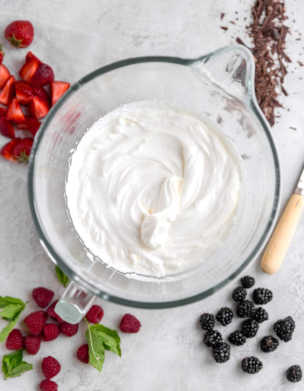 an overhead view of fresh whipped cream in a large glass mixing bowl with raspberries, blackberries, mint leaves, chopped strawberries, and shaved chocolate around it