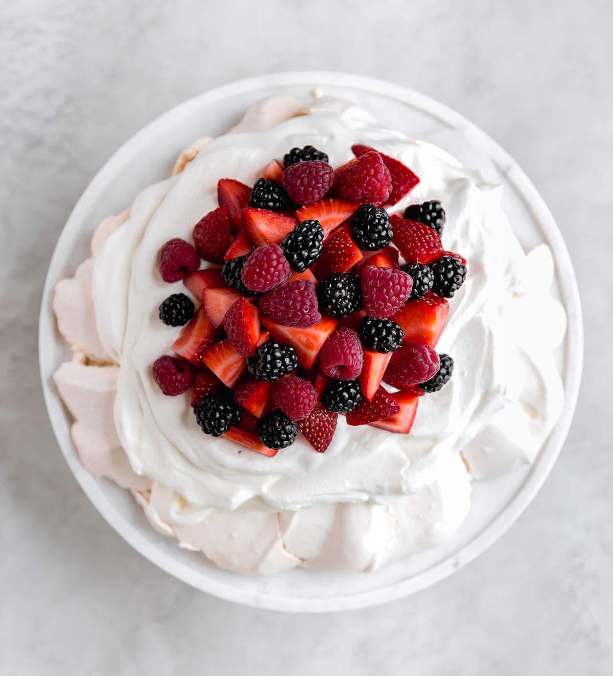 An overhead view of this classic Pavlova recipe served on a glass cake stand with whipped cream and berries.