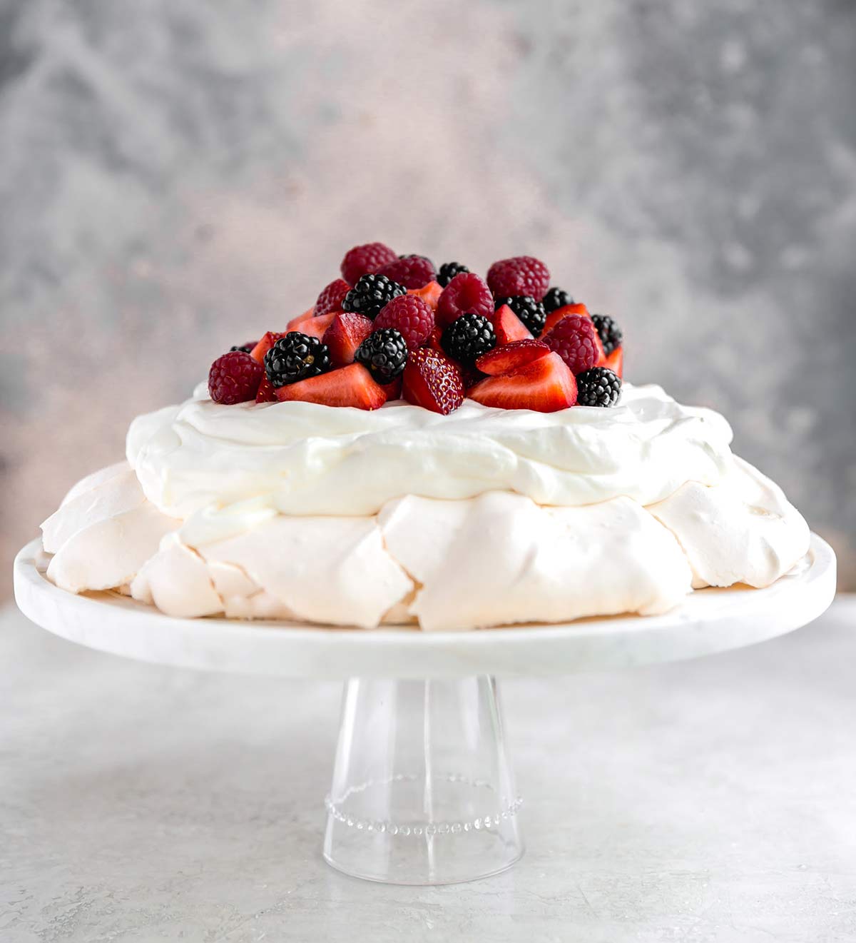 A side view of this classic Pavlova recipe served on a glass cake stand with whipped cream and berries.