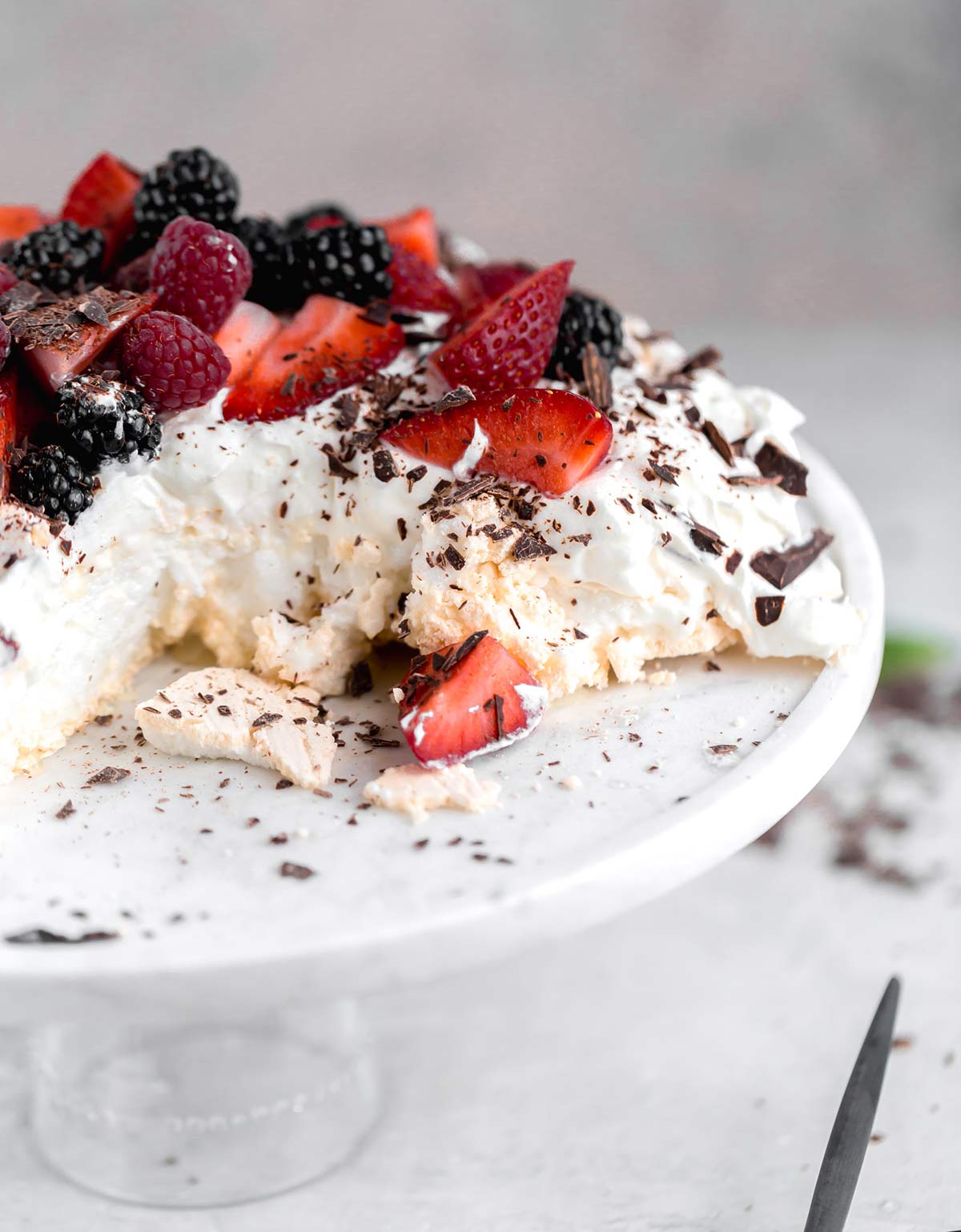 A side view of this classic Pavlova recipe served on a glass cake stand with whipped cream and berries with a sliced removed