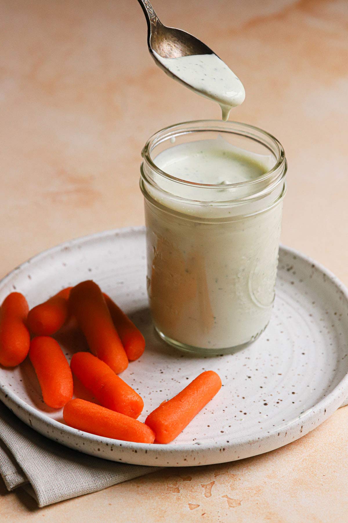 Spoon dipped into jar of homemade ranch dressing on a plate with baby carrots.
