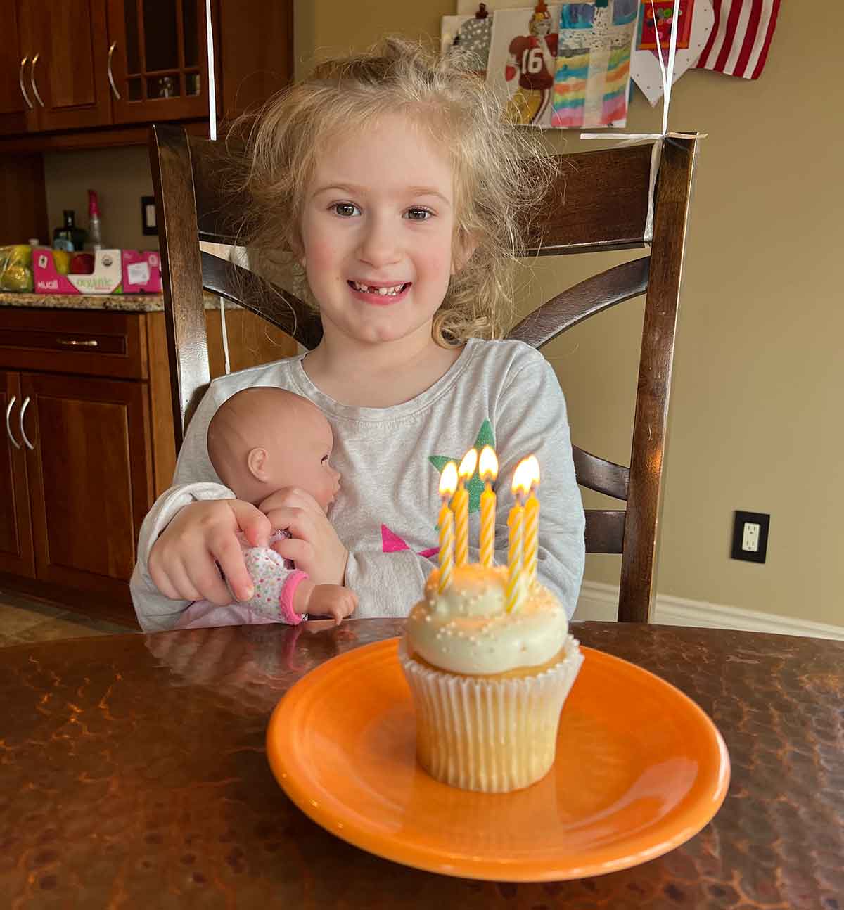 Girl holding a baby doll looking at a cupcake with five candles on an orange plate.
