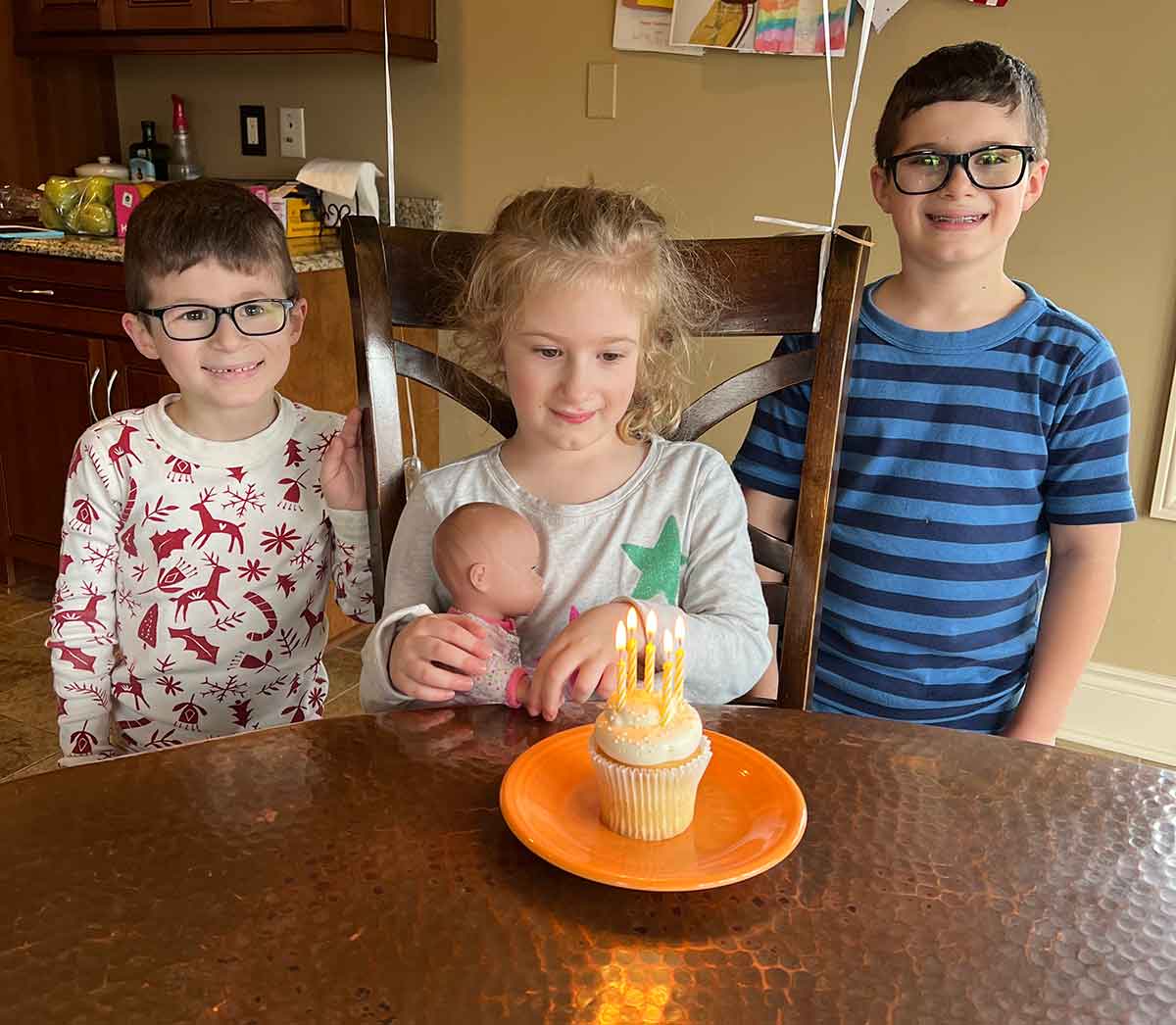 Two boys standing on either side of a girl with a baby doll, preparing to blow out birthday candles.