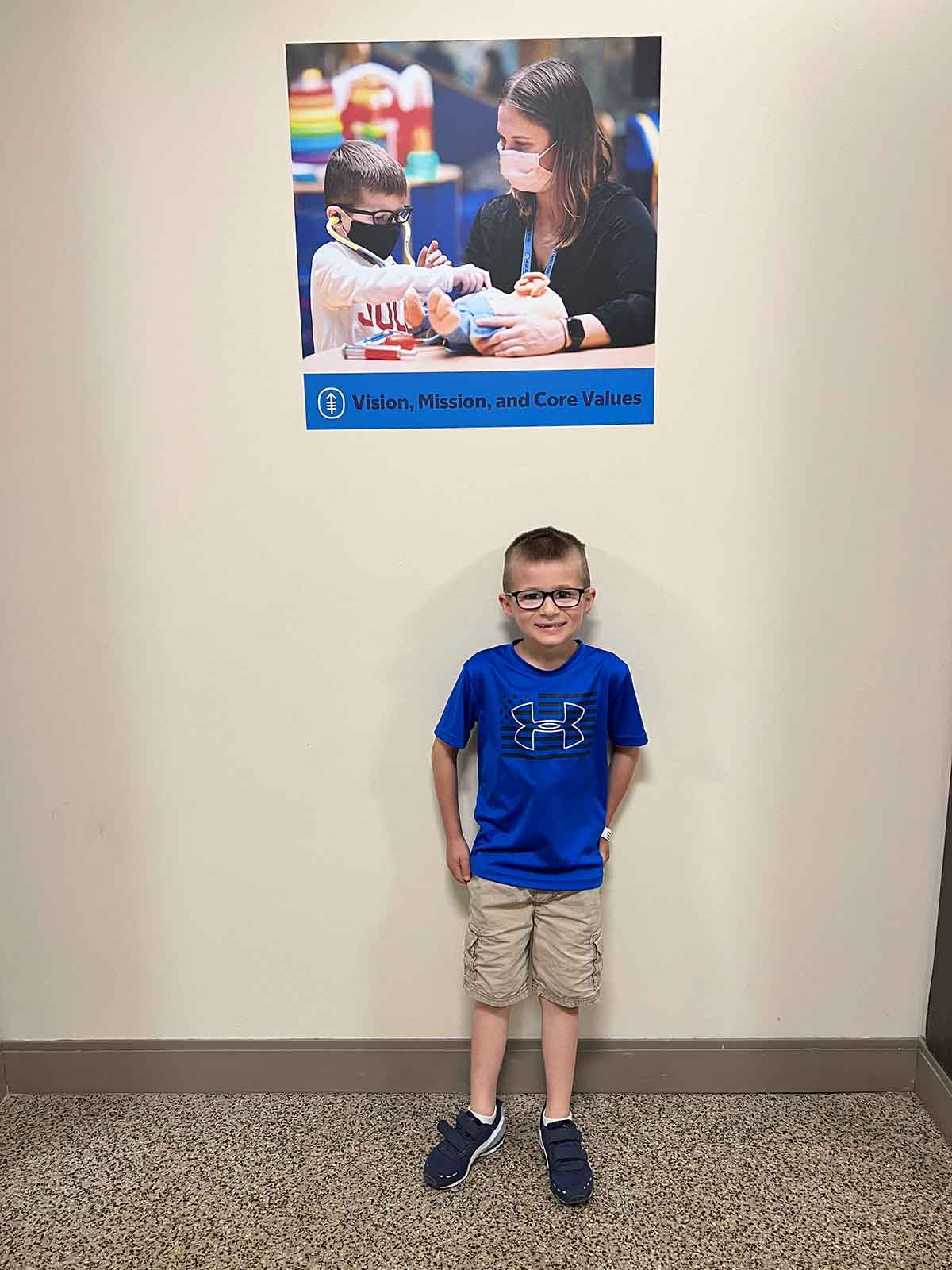 Boy in blue tshirt and khaki shorts standing against a wall below a photo of boy in mask using a stethescope.