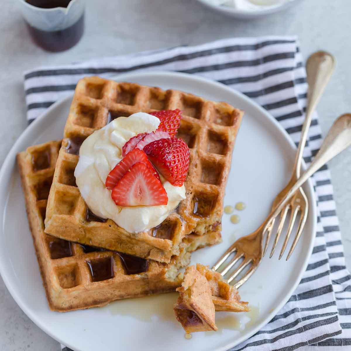 Two buttermilk waffles with whipped cream. strawberries, and maple syrup on top.