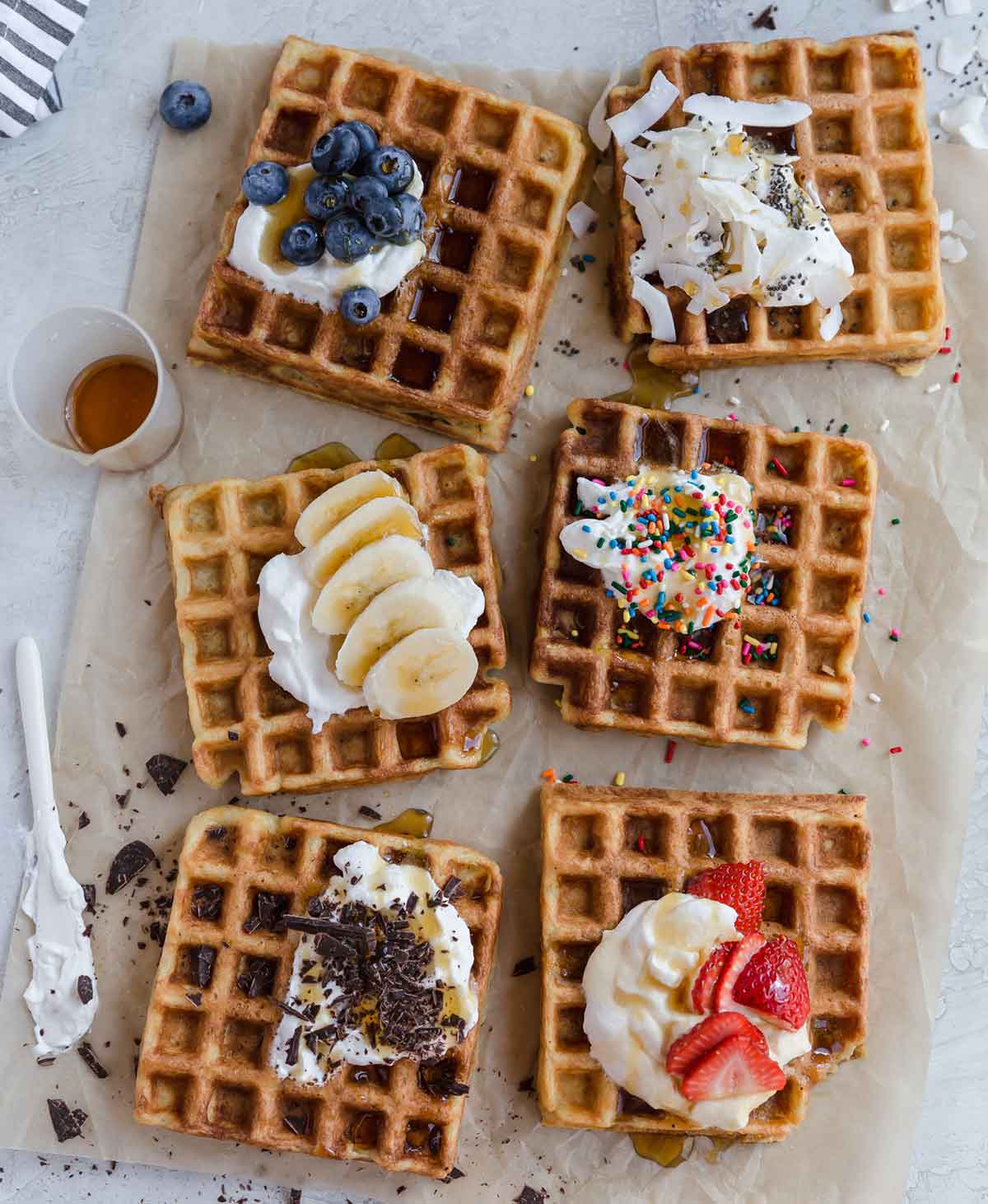 Six square waffles on parchment paper, each with different toppings including whipped cream, fruit, coconut, and sprinkles.