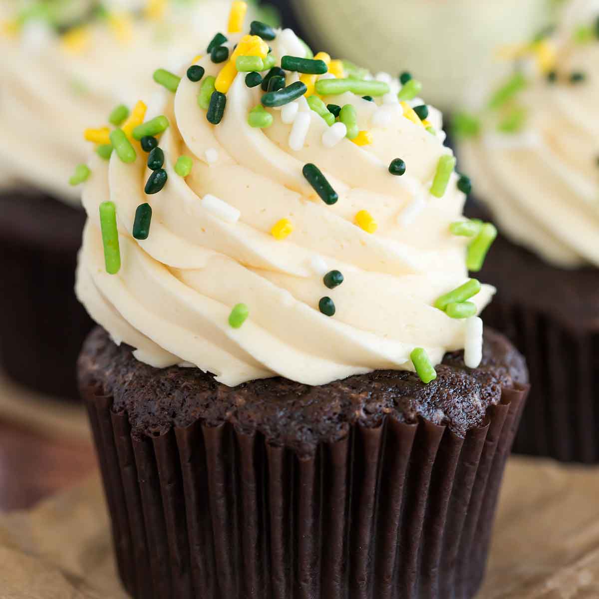 Guinness cupcakes with whiskey ganache filling and Irish cream frosting on brown parchment paper and green sprinkles on top.