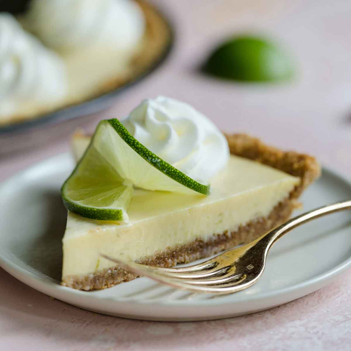 Slice of key lime pie on a plate with a fork, topped with small swirl of whipped cream and lime slice.