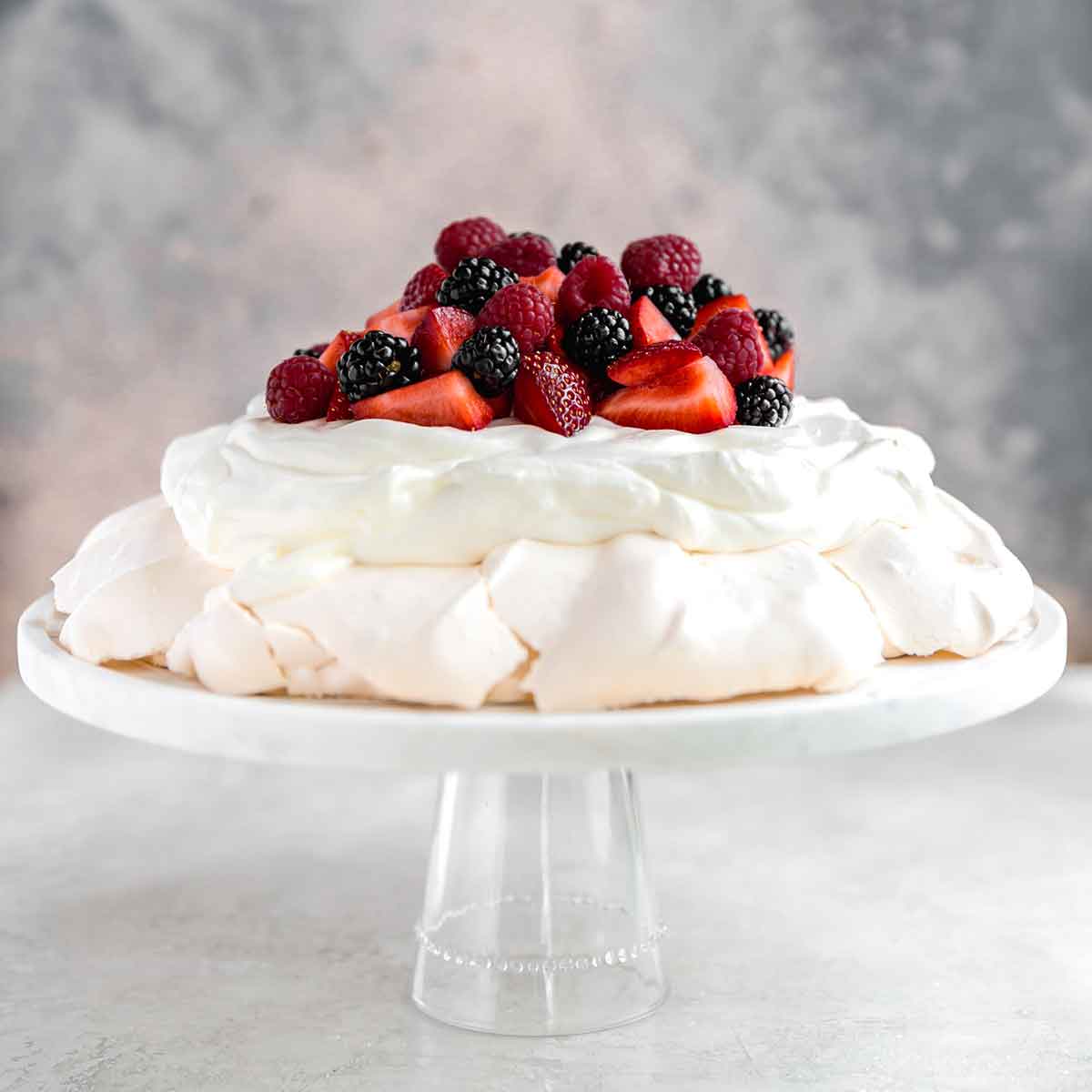 Pavlova topped with whipped cream and fresh berries, on a serving plate.