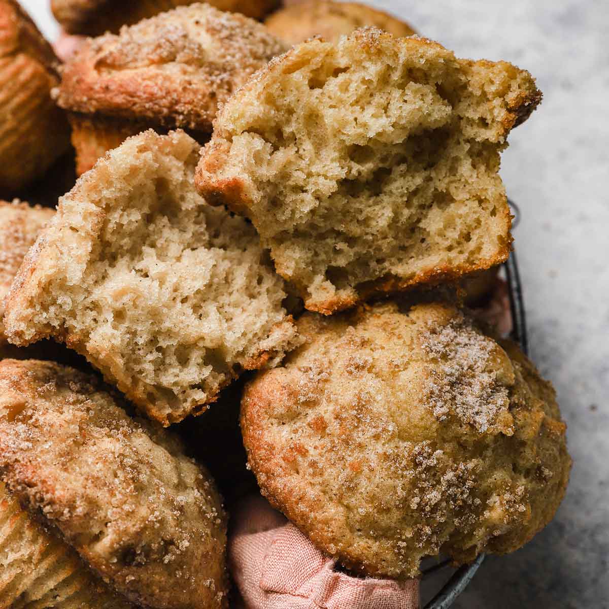 Apple cinnamon muffins in a basket lined with a pink napkin. One muffin is broken in half on top.