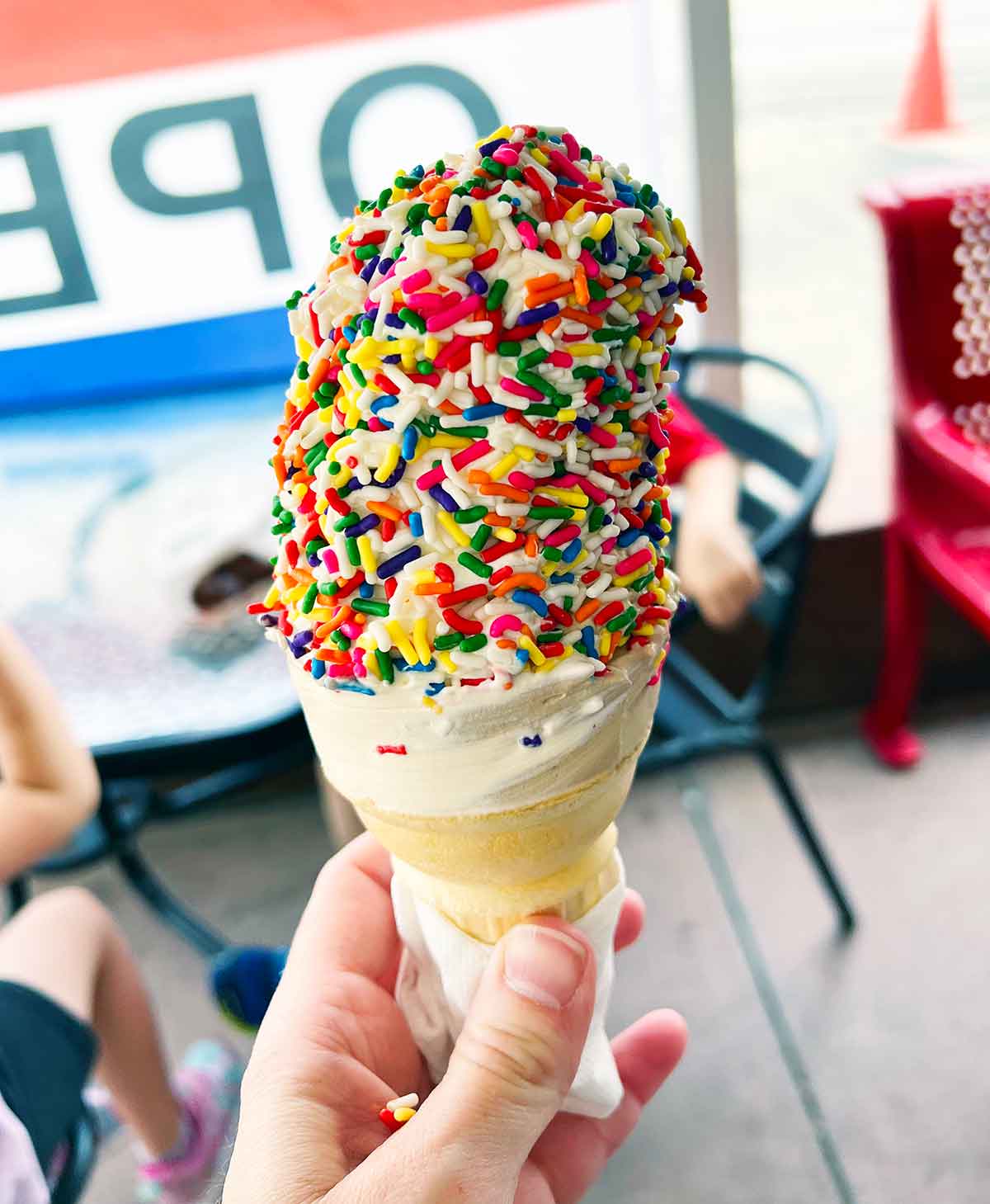 Hand holding a soft serve ice cream cone with rainbow sprinkles.