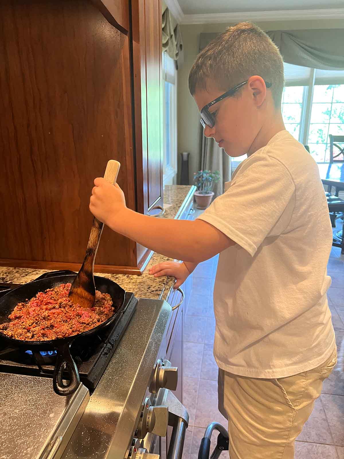 Boy standing at a stove cooking ground beef with a wooden spoon.
