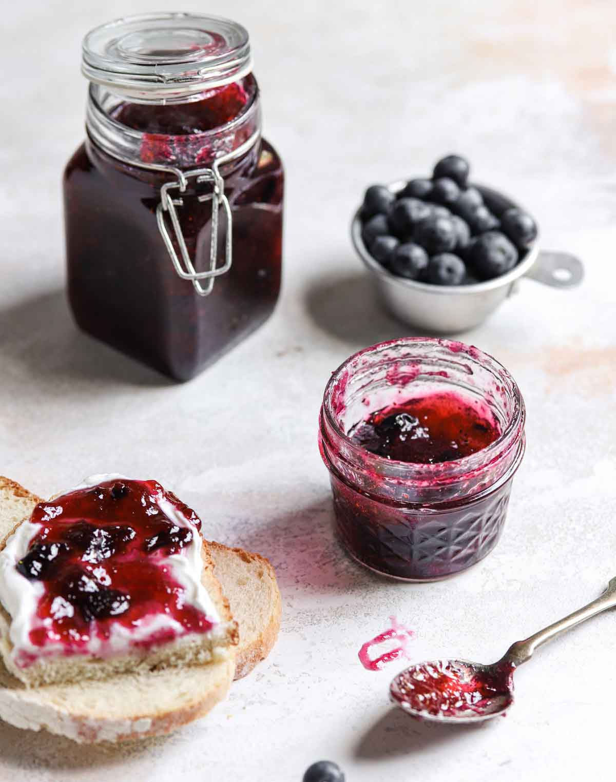 Jar of blueberry jam open with a dirty spoon, bowl of blueberries, and toast with cream cheese and jam around it.