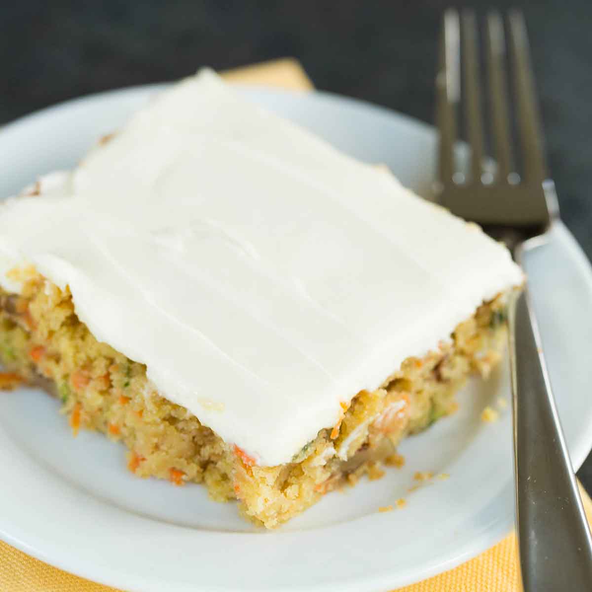 A slice of carrot zucchini bars with cream cheese icing on top on a white plate with a fork to the side.