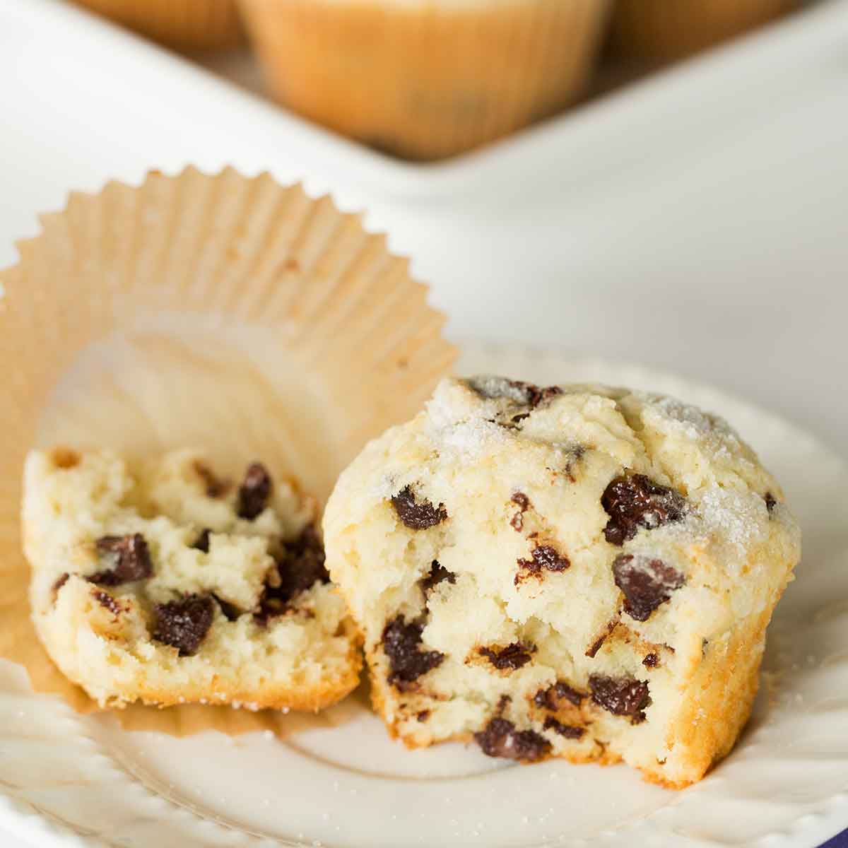A chocolate chip muffins broken in half on a white plate with the liner on the plate.