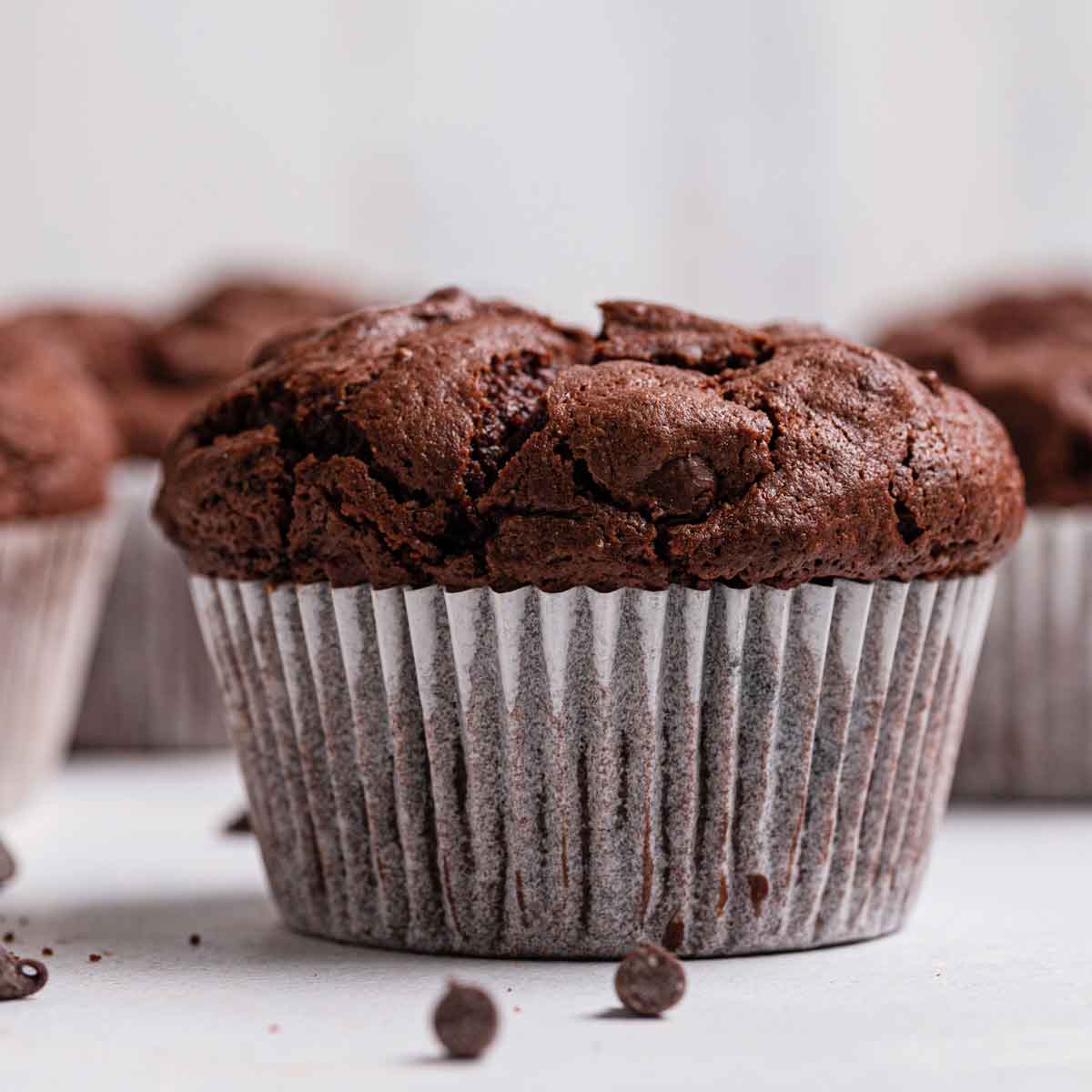 A triple chocolate muffin sitting on a counter in a white liner, with more muffins in the background.