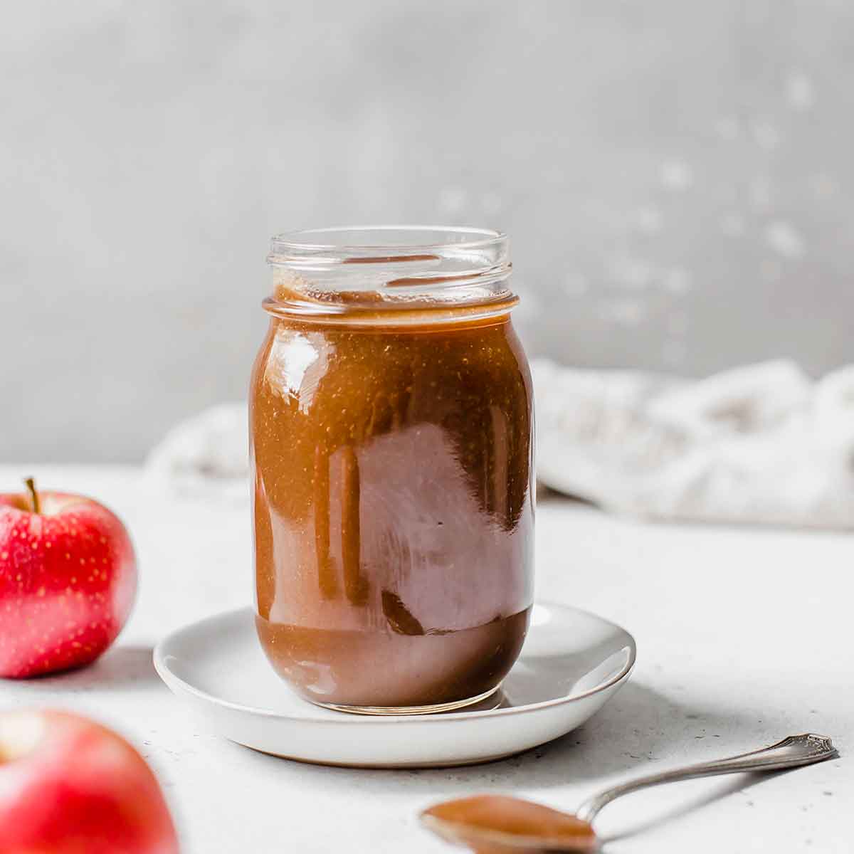 Jar of apple butter on a white saucer with a spoonful of apple butter in the foreground and two apples next to the jar.