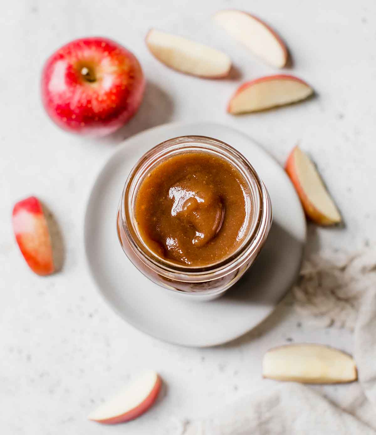 Overhead photo of jar of apple butter on a white plate with sliced apples around it.