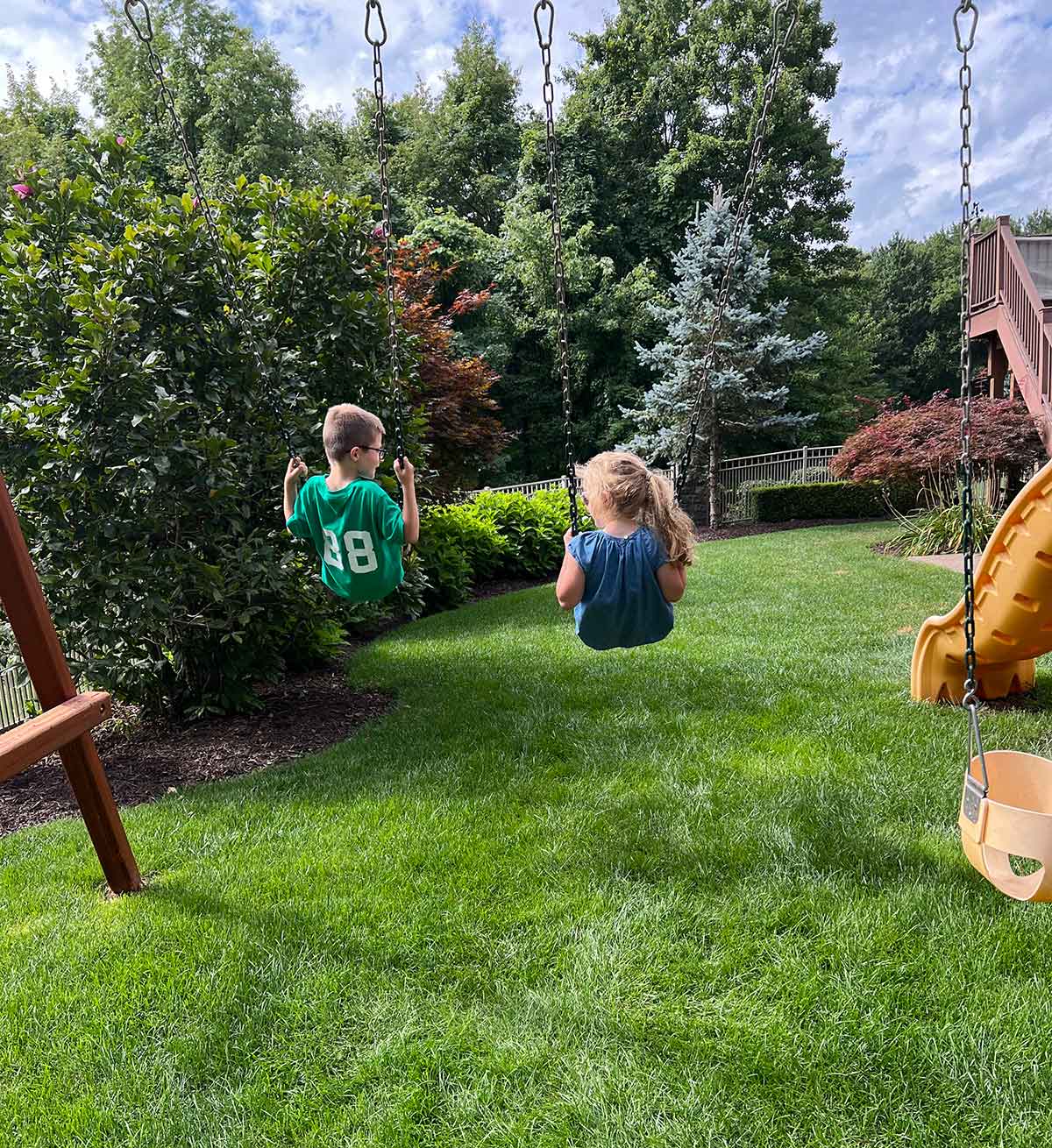 Girl and boy swinging on swings in a backyard with trees in the background.