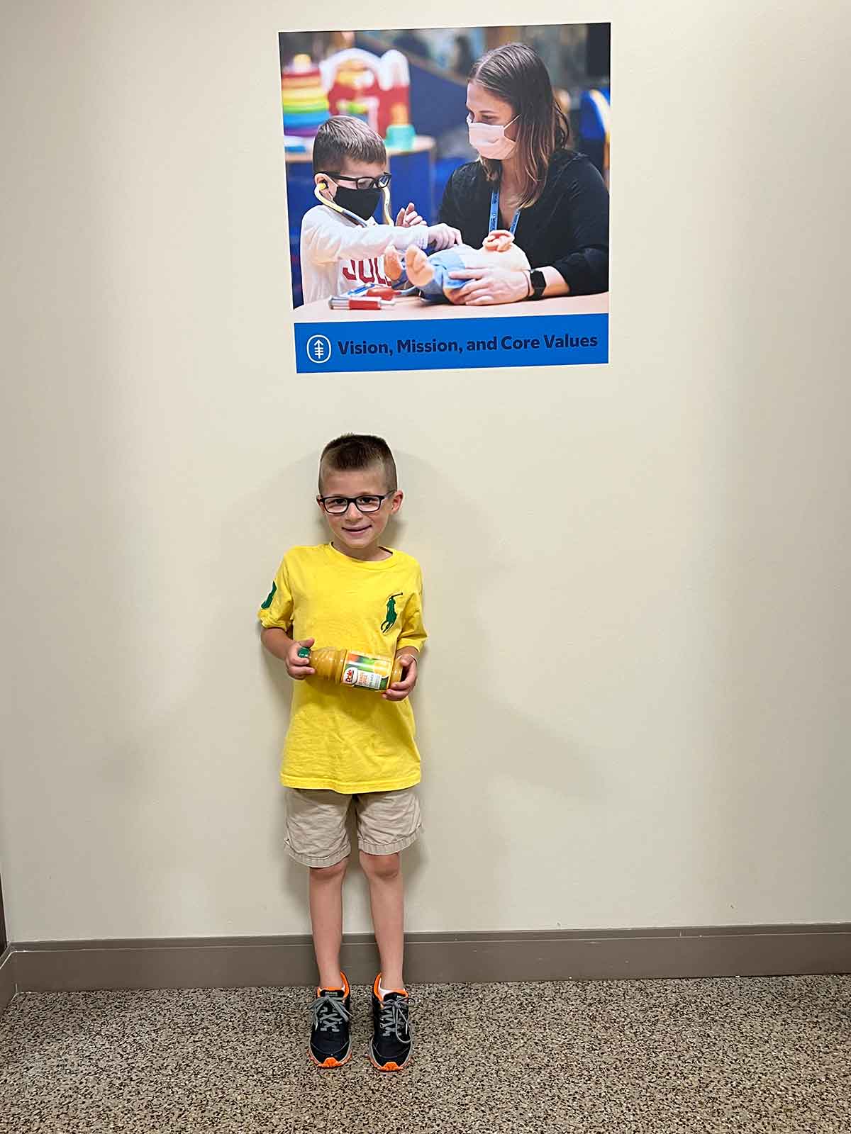 Boy standing in a hallway underneath a photo of a boy using a doctor's kit on a stuffed animal.