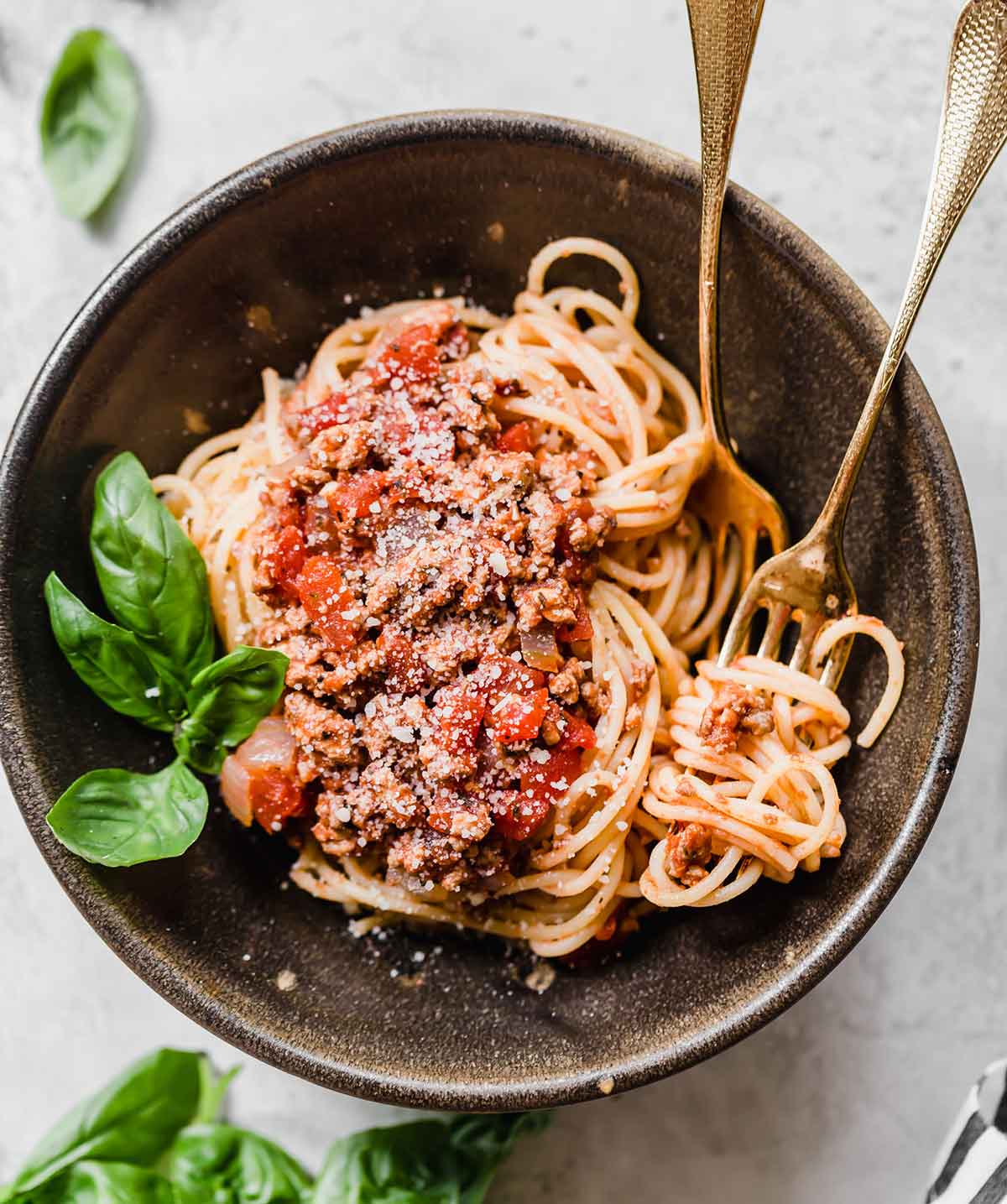 A bowl of spaghetti with meat sauce and two forks and a sprig of fresh basil in the bowl.