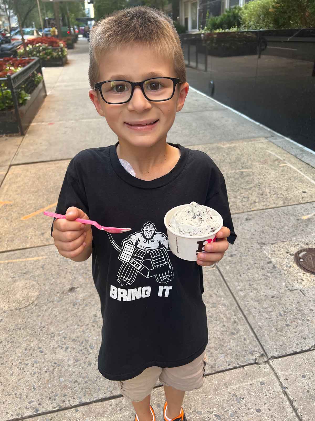 Little boy standing on a city sidewalk holding a cup of ice cream.