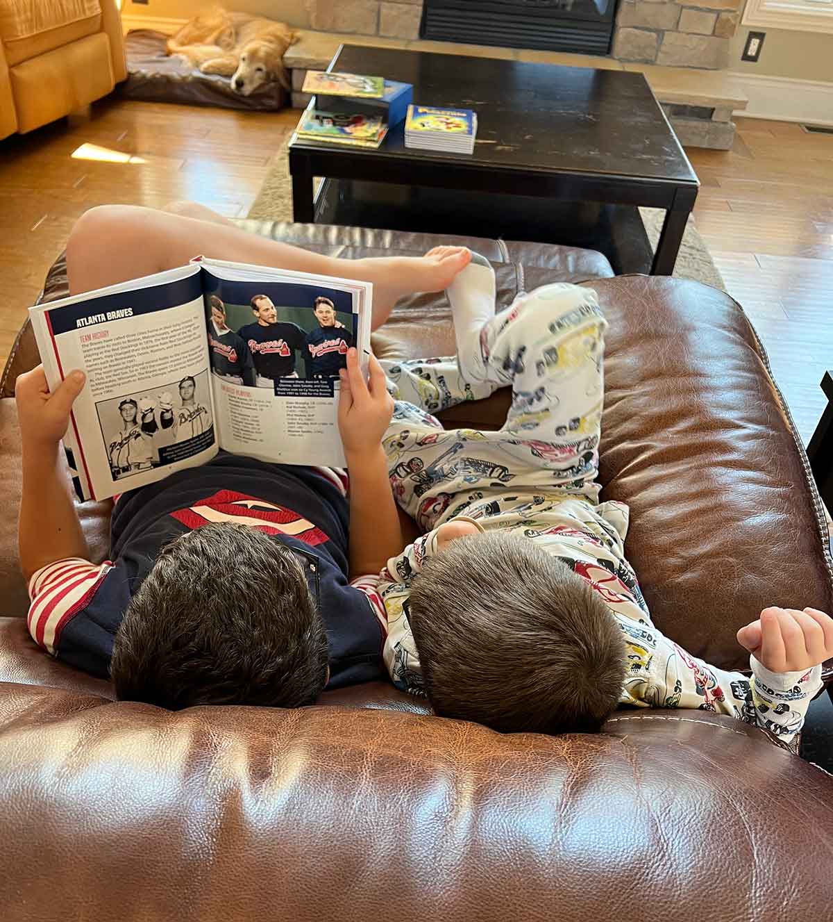 Two boys sitting next to each other on a couch reading a baseball magazine.