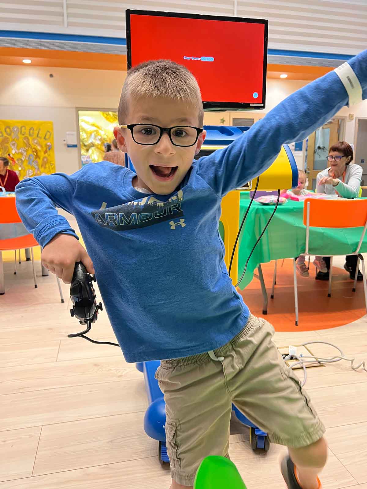 Boy in a blue shirt and black glasses holding a video game controller standing on one leg and smiling.