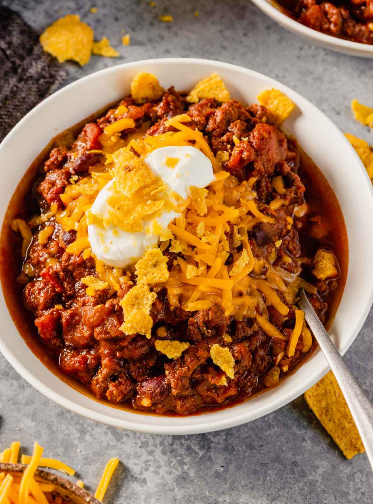A close up photo of beef chili in a white bowl topped with shredded cheese, sour cream, and corn chips.