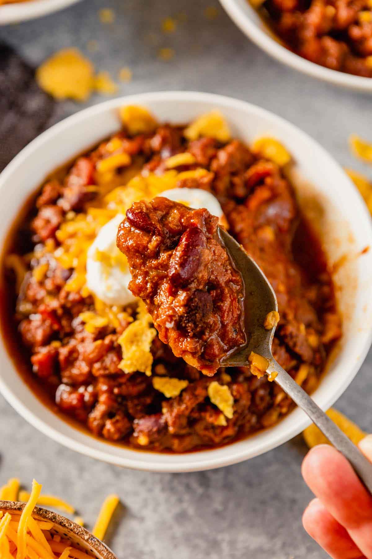 Close up photo of spoonful of chili held over a whole bowl of chili with sour cream and corn chips visible.