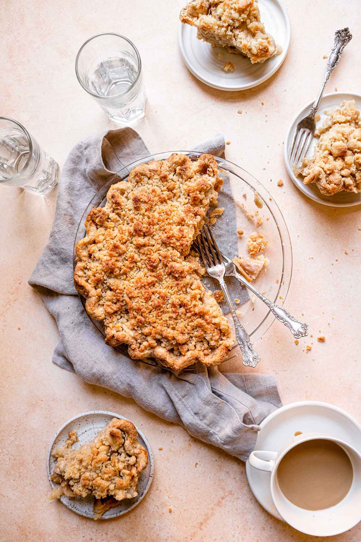 Overhead photo of Dutch apple pie in glass pie plate, with slices removed and on small white dishes in the surrounding area.