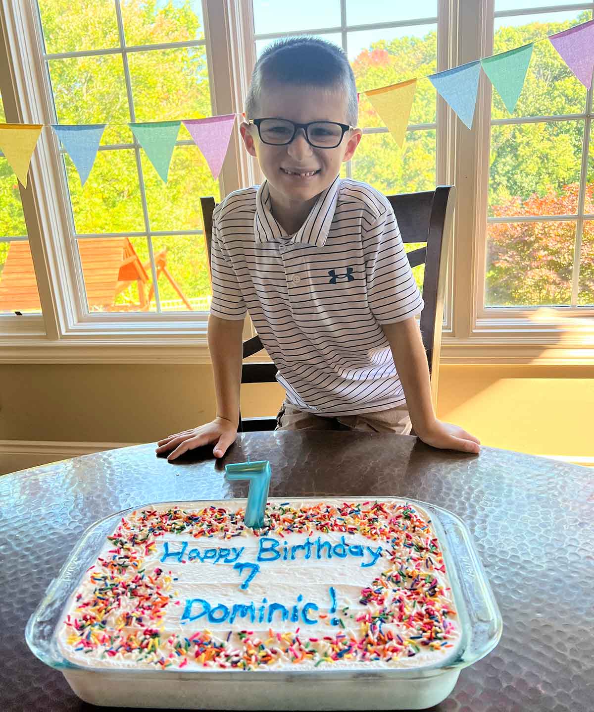 Little boy in a striped polo shirt smiling over a birthday cake with a number seven candle in it.