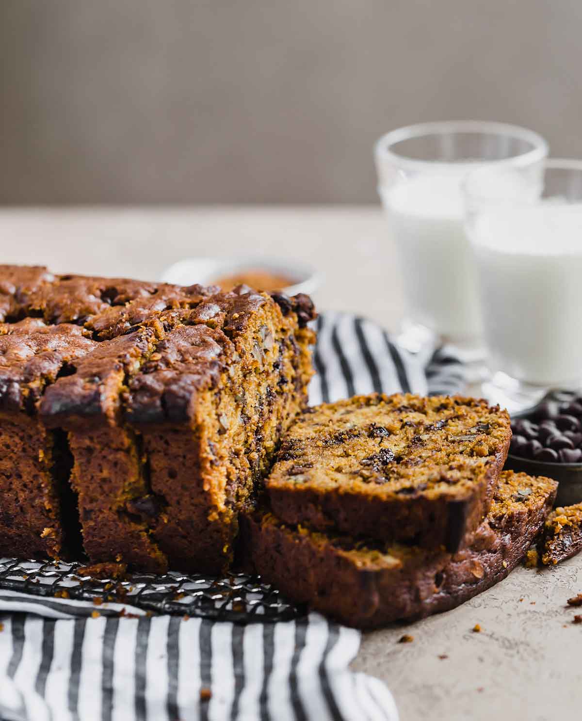 Pumpkin bread with chocolate chips and pecans sliced on a tea towel with glasses of milk in the background.
