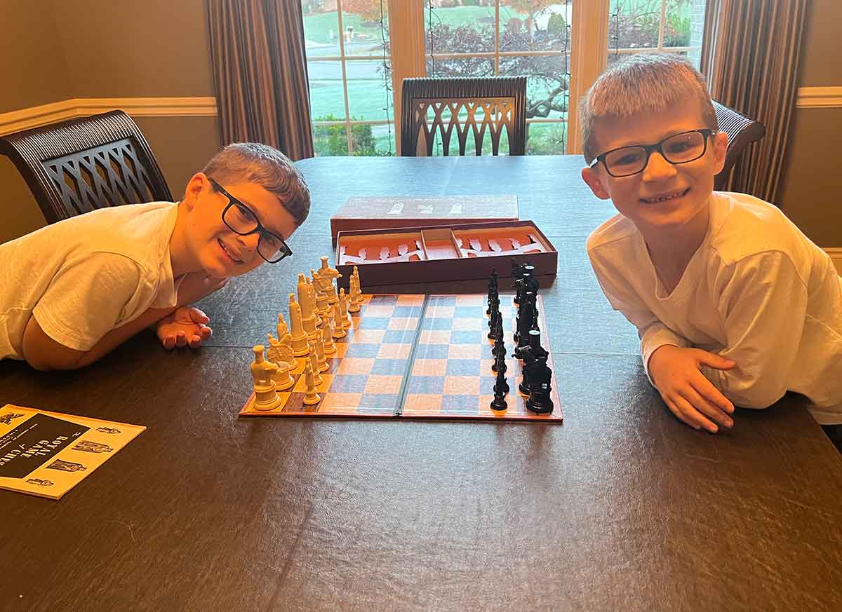 Two boys sitting on opposite sides of a chess board at a dining room table, smiling at the camera.