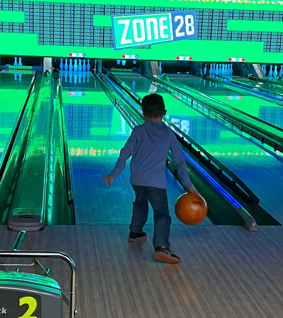 Little boy throwing an orange bowling ball, bright green and blue lights in background.